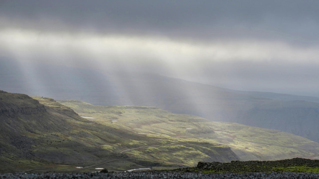 A colour photo of a landscape with a canopy of heavy cloud at the top of the shot. The foreground is the edge of a road with a sheer drop down to a small plateau in shadow. A dirt road can be seen going across it. Beyond that is another plateau with greenery and rocks highlighted by strong sunbeams emanating from the top of the photo.