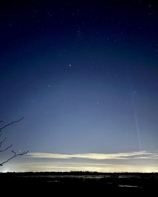 A wide view of a starry sky over a rural scene with puddles of water reflecting the band of light pollution at the horizon. A beam of light points up from the horizon into a cluster of stars.