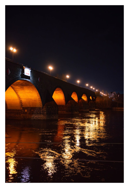 Foto im Hochformat. Nachts. Blick entlang einer Steinbrücke, die übers Wasser geht, aus Höhe der Wasseroberfläche fotografiert. Unter der Brücke sind viele Bögen, durch die Schiffe fahren können. Diese sind jeweils beleuchtet. Auf der Brücke sind Straßenlaternen. Das Licht spiegelt sich im dunklen Wasser.