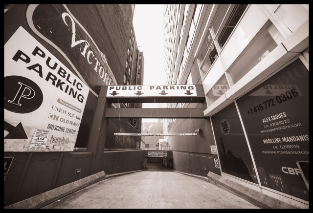 A brown-toned, monochrome photograph of a driveway wedged between two tall buildings in downtown San Francisco. Taken with an ultrawide lens, there's interesting distortion in the photograph as the buildings bend inward overhead while reaching out on the sides to wrap around the photographer.

Anyway, what you see here is a driveway dropping down into a basement level parking lot. There are large signs on the left, overhead, and further down the driveway, with arrows, announcing Public Parking with monthly rates available. Higher up on the left wall of one of the buildings, there's an old ghost sign for Victoria or Victorian... not sure. Also overhead, there's a narrow slice of sky visible, glaring while iit casts difuse light onto the driveway below.

Not counting Google Streetview, this is possibly the first photograph ever taken of this parking lot entry. I am proud.