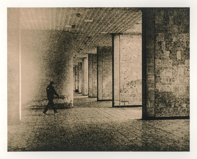 This lith print shows a stark, atmospheric urban interior — a long concrete corridor supported by massive rectangular columns that recede rhythmically into the distance. The space feels cavernous and slightly oppressive, emphasized by the heavy grain, mottled textures, and uneven tonal transitions typical of the lith printing process.

A solitary human figure appears blurred in motion on the left side of the frame, walking through the corridor. The figure’s indistinct outline suggests transience and anonymity, as if the person is momentarily passing through an indifferent architectural landscape. The motion blur contrasts with the rigid, immovable geometry of the columns and tiled floor.

The surface of the print is alive with lith grain: speckled shadows, rough highlights, and cloudy midtones that give the image a weathered, almost decaying quality. This texture softens the hard lines of the architecture while adding a sense of time, erosion, and instability.

Overall, the image evokes themes of isolation, movement through liminal space, and the tension between human presence and brutalist architecture.