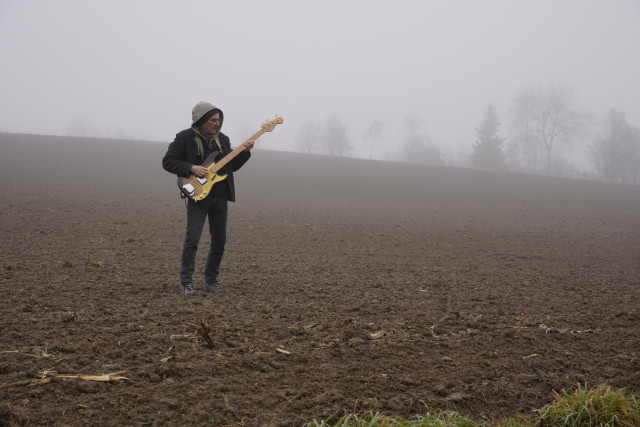 Ein Bassist steht an einem nebligen Nachmittag auf einem Acker und spielt Bass.