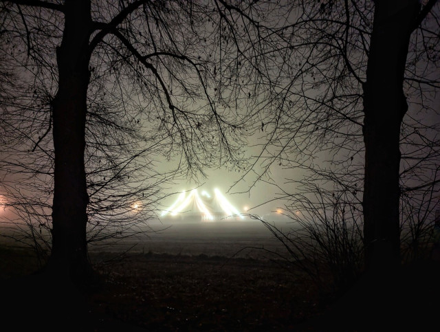 This image captures a misty scene framed by the silhouetted branches of leafless trees. In the distance, the bright, vertical lights of an illuminated circus tent pierce through the fog, creating a striking and mysterious focal point. The tent’s lights form a triangular shape, adding depth and intrigue to the composition. The overall atmosphere is serene yet slightly surreal, blending the natural darkness of the trees with the artificial glow of the circus tent.