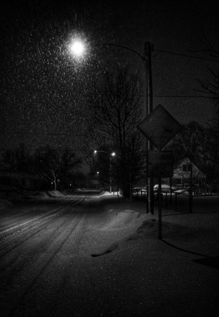 Looking down a dark residential road at night as the snow falls. Snow covers the road and the ground, houses can be seen on both sides of the road as well as a few trees. A light post with a bright light highlighting the falling snow is seen in the foreground as well as another sign.