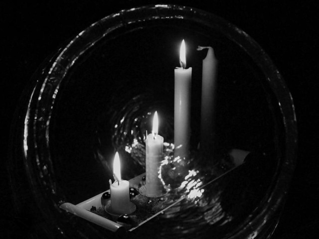 An Advent candlestick with three lighted candles seen slightly obliquely from the side, with the first candle at the front. Around it all, a faint ring of light and glare is visible in the center of the image.