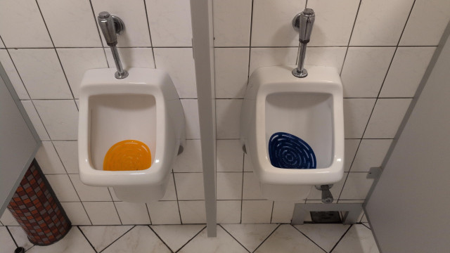 Foto von zwei Pissoiren. Im linken ein ovaler, orangener, im rechten ein blauer Einleger (Urinalsieb).