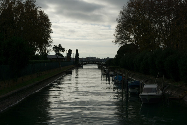 Clouds filter pale sunlight, reflected silver in the smooth undulanting surface of a canal. Boats line one edge, half-hidden in the shadows of silhouetted trees. A bridge crosses in the distance.