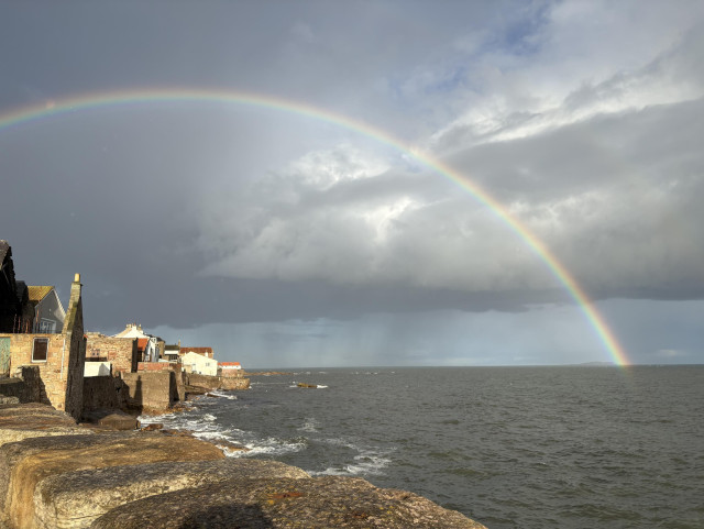 A rainbow arcs out over brightly illuminated coastal buildings and a rocky shoreline and plunges into a rough grey sea. The sky in the distance is ominous and stormy, lending the rainbow and buildings a luminous quality. 
