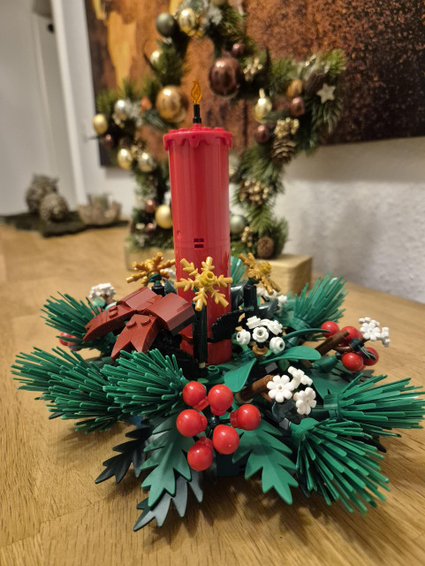 This image features a festive holiday centerpiece made from building blocks, designed to resemble a traditional Christmas decoration. At the center of the arrangement is a red candle, surrounded by green pine branches, red berries, white flowers, and gold snowflakes—all constructed from colorful blocks.

The centerpiece is placed on a wooden surface, with a decorated Christmas tree in the background, adding to the holiday ambiance. The tree is adorned with ornaments and garlands, enhancing the festive atmosphere of the scene. The overall arrangement is charming and creative, capturing the spirit of the holiday season in a playful and unique way.