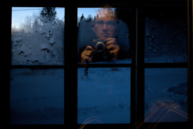 The view through two sets of windows, over the sink and across the verandah and out into the snow. A yellowish light illuminates me in one pane. At the lower right ghostly cutting boards glow in a similar fashion. There is frost on the outer windows. The sun is rising to the left.