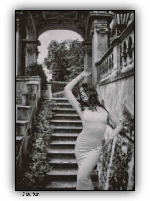A black-and-white analog photo shows a woman in a long, fitted dress posing gracefully at the foot of grand stone steps. The model is my wife. Her posture is upright, arms placed elegantly, gaze directed beyond the frame. The dress hugs her figure, highlighting the line of her hips and shoulders, with the fabric catching the light in a sculptural way. Behind her, the steps lead up to a classical garden pavilion with arches and columns, resembling a small temple. Flower pots line the stairs, adding softness to the symmetry. On the right side there’s a stone balustrade and wall, with lush greenery stretching beyond. The whole scene radiates calm, elegance, and a touch of theatrical presence.
