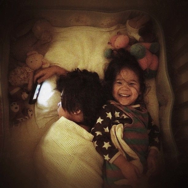 2 siblings in one bed, the 5-year old boy is watching a phone screen, his 2-year old sister is smiling towards the camera.
