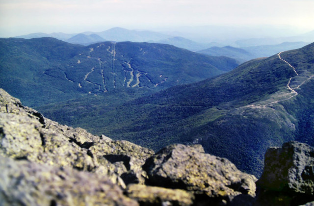 Beneath a clear, bright very pale blue sky, everything in this pic is bathed in sunshine. We are standing on a jagged outcrop of light gray granite covered with pale green and dark green lichen high atop a mountain. To our right are 2 massive ridges running down from a lofty mountain just offscreen right. Their flanks are wooded, their rocky crests are open and covered with stunted dark green alpine vegetation which extends a short distance down their sides before gradually giving way to trees. Several trails are visible as meandering light gray strands. Beyond this mountain on the left are a series of wooded green mountain ranges cascading to he misty horizons. A massive spiderweb of intersecting ski runs stands out along the face of the closest of these mountains.