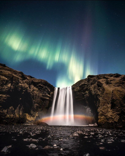 A beautiful photograph of a waterfall dropping into a pond at the base of a rocky cliff. The moonlight is creating a moonbow in the mist of the waterfall. Above, the night sky shimmers with the green and purple waves of the aurora borealis.