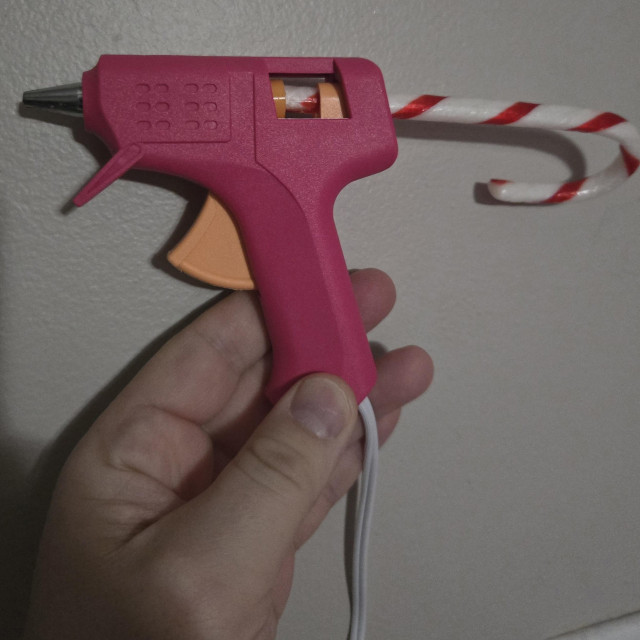 Photograph of a white person's hand holding a pink hot glue gun against a light colored wall with a candy cane in the back of it.