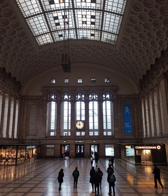 Wide interior of Leipzig train station hall with a high arched glass ceiling, patterned stone walls, and tall windows. A large clock hangs above the main entrances, while a few people walk across the spacious tiled floor. Shops line the sides of the hall.