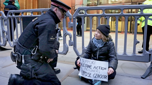 Greta Thunberg sits cross-legged on the ground, holding a handwritten sign that reads "I SUPPORT PALESTINE ACTION PRISONERS" and "I OPPOSE GENOCIDE". A police officer in a black uniform and peaked cap kneels directly in front of her, appearing to speak to her. Behind them are grey metal crowd control barriers and other police officers standing in the background.