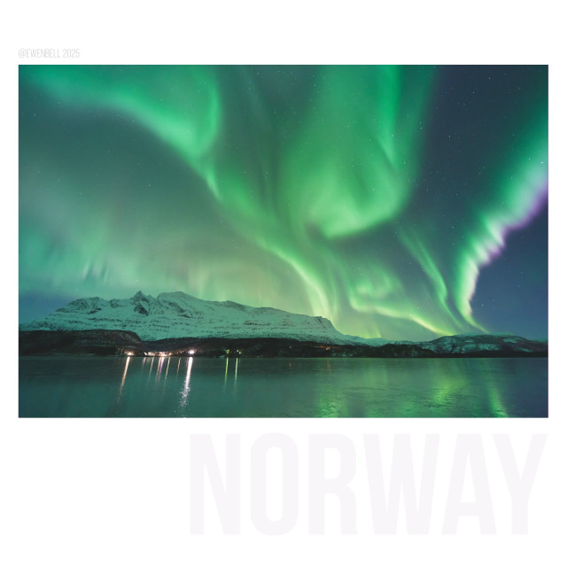 Auroras reach up from the western horizon across the fjord. A few lights from a Sami village reflect across the ice. Christmas in the Arctic, December 23 2025.