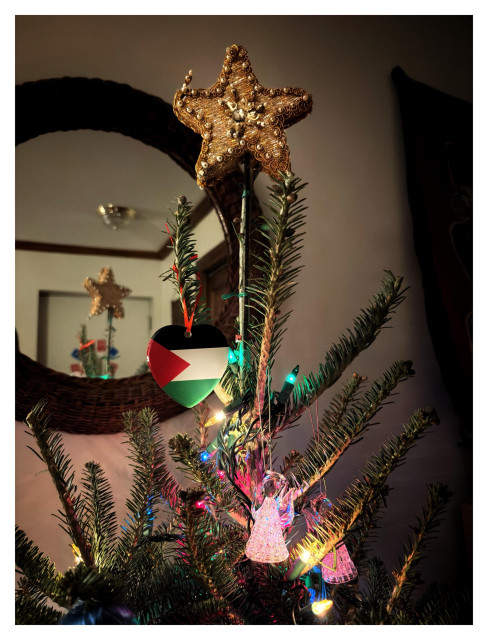 Photo of the upper portion of a Christmas tree, featuring a star made of woven golden fiber and, just below it, a heart-shaped ornament with the design and colors of the Palestinian flag (black, white and green stripes with a red triangular field). Multicolored string lights and glass angels also adorn the branches. Behind the tree is a mirror in a circular frame of woven wood fibers, which reflects the gold star on top of the tree and some of the ceiling and walls behind the camera.