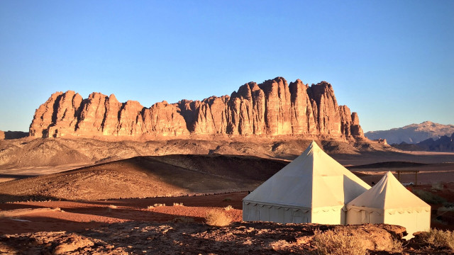 Two white, pyramidally roofed tents stand in Wadi Rum. The sun casts long shadows across the open terrain and illuminates distant sandstone.