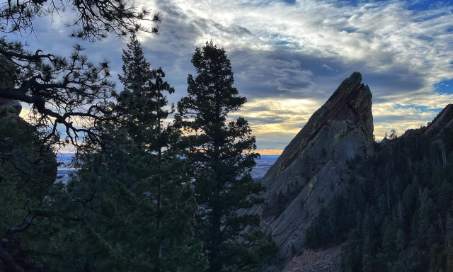 Looking through tall, dark green pine trees toward an uptilted rock formation called the Third Flatiron, with mostly cloudy skies, their edges lined with light from the hidden Sun, in the background.