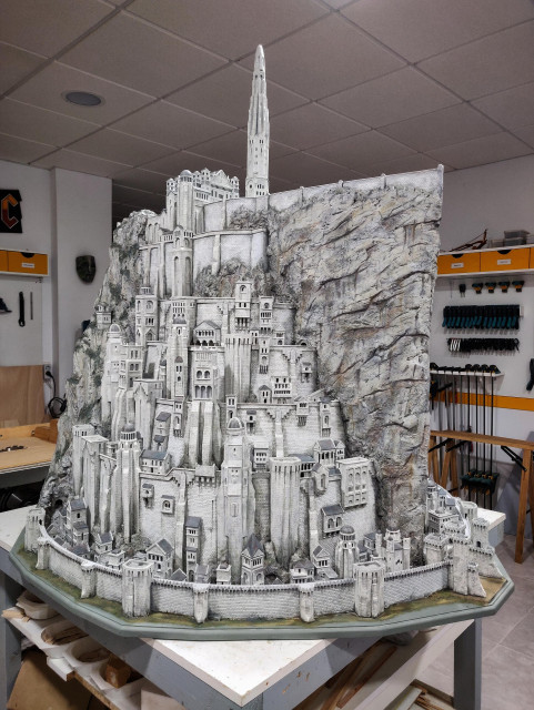 A model of a tall fortress city. Buildings are arranged in different circular levels, each smaller than the one below, like a wedding cake. All levels are crowded with lots of different buildings and towers of various sizes, giving the city a busy look. On the top level there's a palace and a tall spire. There's a huge flat rocky spur slicing the city in half. The buildings and rocks are mostly white. The model rests on a table, with some tools visible in the background.