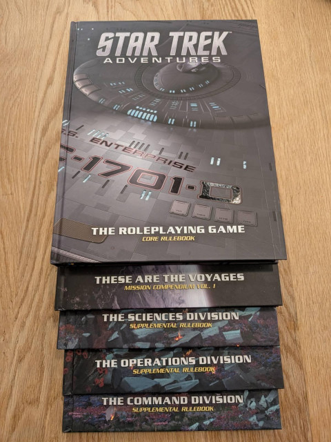 5 books stacked on a wooden table. The top book is Star Trek Adventures The Roleplaying Game Core Rulebook. The four other books only shown their titles which are These Are The Voyages Mission Collection volume 1, then three supplemental rulebooks, The Science Division, The Operations Division, and The Command Division.
