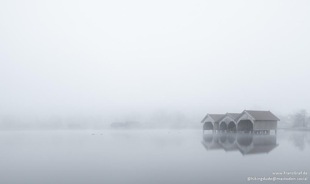 This image captures a serene and almost mystical winter scene. In the center of the frame, there are three connected wooden boathouses or huts, elevated on stilts above a calm body of water. The water is so still that it perfectly reflects the structures, creating a mirror-like effect.

The entire scene is enveloped in a thick fog, which obscures the background and adds a sense of mystery and tranquility. The fog softens the outlines of the boathouses and blends the horizon seamlessly into the sky, creating a minimalist and ethereal atmosphere.

The overall mood of the image is calm and peaceful, evoking a sense of solitude and stillness. The muted colors and the foggy conditions contribute to the timeless and tranquil ambiance, capturing the quiet beauty of a foggy winter morning by the water.