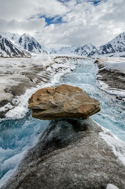 A wide meltwater stream flowing across the surface of a glacier, with bright blue water cutting through ice and compacted snow. A large, flat-topped boulder sits balanced on a narrow ridge of ice in the foreground, dividing the channel. Snow-covered mountains rise on both sides of the valley under a cloudy sky.