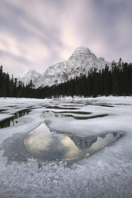 A snowy winter scene taken ontop thin ice. Holes in the ice reflect the sunrise light and the snowy peak in the distance. The ice over the river is covered in thin ice with frost ontop.