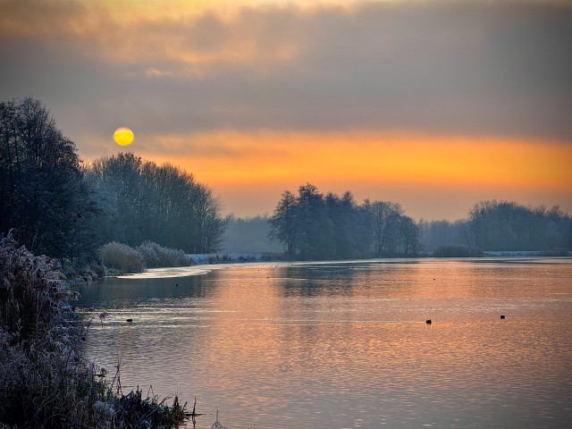 A serene winter landscape featuring a calm river reflecting a vibrant sunrise. Frost-covered trees line the banks, while a soft mist hovers above the water. The sky transitions from warm orange to muted gray, creating a peaceful atmosphere.