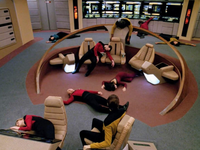 Star Trek The Next Generation scene. The bridge of the Enterprise is shown from above looking down. Data sits at his console looking back at the rest of the crew, who are all passed out in various situations; on the floor, slumped over a console, or half falling off their chairs. Worf, notorious Klingon warrior, dangles over the tactical station. No caption. 