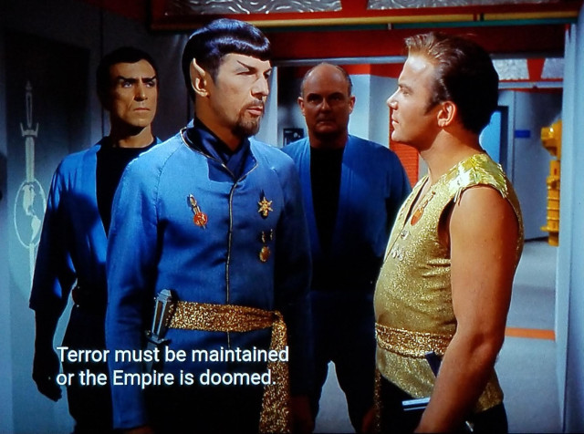 Star Trek The Original Series scene. We're in a corridor on the Enterprise. Two non series regular crew members are standing behind Spock in a bodyguard stance. (Tough guy bodyguard vibe, not Kevin Coster/Whitney Houston bodyguard vibe). Spock is wearing a dress uniform with extra flair, medals, and pins, a golden sash, and has a goatee. He's looking at Kirk, who seems a bit cocky and is wearing a glittery fancy gold sleeveless uniform. Closed caption reads, "Terror must be maintained or the empire is doomed."