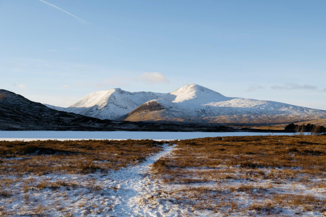 A frozen lochan in the foreground with a narrow snowy path leading towards it, backed by the broad, snow dusted hills of the Black Mount under a clear blue winter sky.