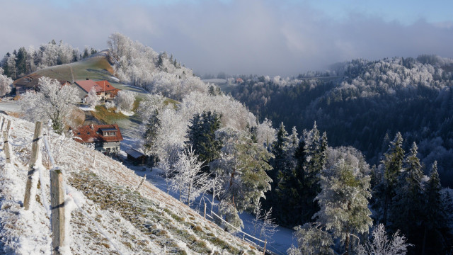 A winter landscape featuring snow-covered hills, frosted trees, and quaint houses with red roofs nestled in a valley. The scene is bright with blue skies and scattered clouds.