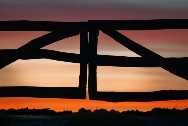 Gegen orangefarben marmorierten Himmel die Silhouette eines Holztores im Stile eines Ranchzauns. 
Abstrakt durch den Blickwinkel.