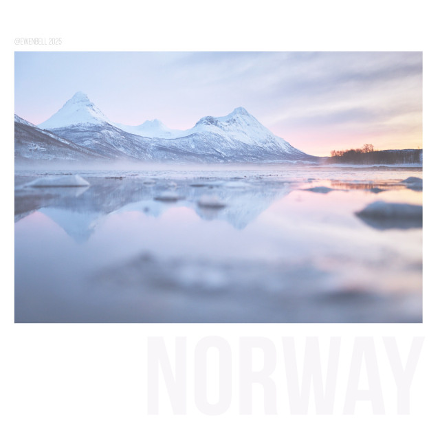 Distant mountains rise above the fjord, reflected in the calm waters, Snow covers everything. A hint of warm light has escaped the horizon to the south.
