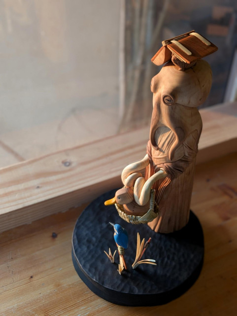 Woodcarved scene on a round base. An humanoid octopus wearing a dress is holding a basket with a duck inside of it. On the head of the octopus, a grumpy frog taking shelter of a big ancient book. Next to them all, a sword is coming out of water among bulrush. On the top of it sits a kingfisher.