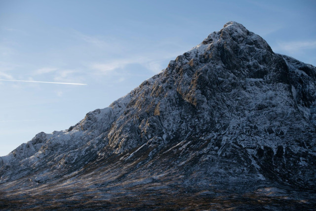 A winter mountain rises steeply from a broad, open glen, its rocky face dusted with snow and catching cold blue light. The foreground is dark, rough moorland, while the sky above is pale and clear, crossed by a thin white aircraft contrail. The scene feels stark, expansive and quiet, with no people or buildings visible.