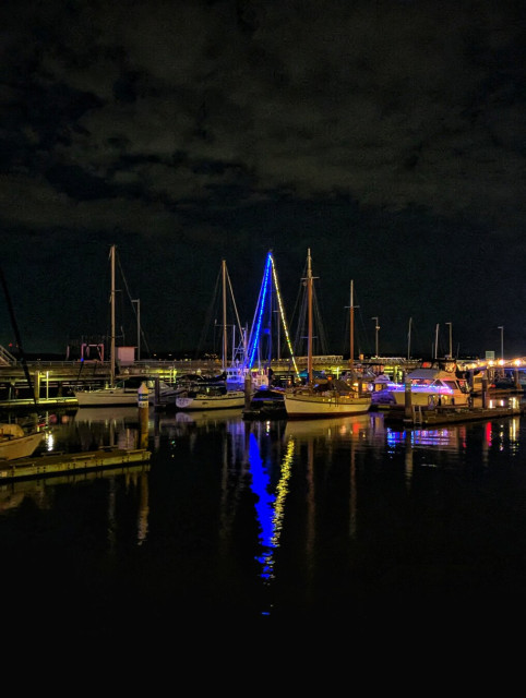 This nighttime photograph captures a serene marina scene. Several sailboats and yachts are docked along wooden piers, their masts reaching up into the dark sky. One of the boats is adorned with vibrant blue and yellow LED lights, creating a striking reflection on the calm, dark water below. The lights from the boat and the marina cast a warm glow, illuminating parts of the pier and the surrounding boats. The sky is overcast, adding a moody atmosphere to the tranquil setting.