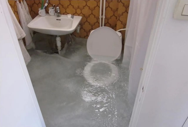 An old style toilet and sink where the pipes are exposed. There has been a leak at some point, and the room filled up with water to just above the toilet seat, but everything is frozen so the toilet seat can now be seen inside a block of mostly clear ice.