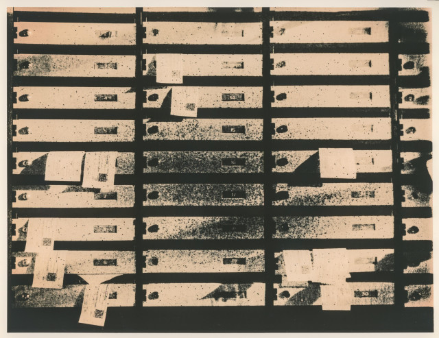 This image shows a wall of metal mailboxes arranged in a strict horizontal and vertical grid inside the lobby of a residential building. Each mailbox is narrow and rectangular, with small number plates and keyholes, some marked by smudges, scratches, and traces of wear. Several slots are partially covered with folded pieces of paper, giving the surface an improvised, slightly chaotic look.

The high-contrast, grainy texture and muted tones emphasize repetition and age, turning the functional mailboxes into an almost abstract pattern of lines, rectangles, and dark gaps