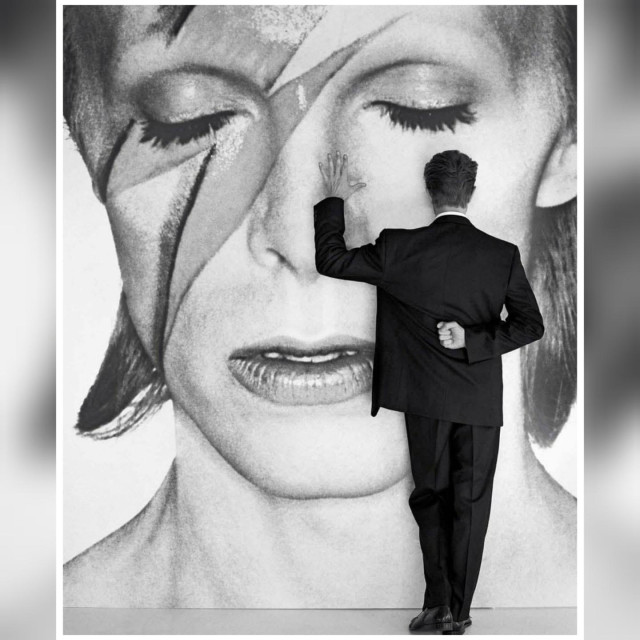 Black and white picture of Bowie wearing a black suit, having his back turned to the camera as he is facing a wall with a large picture of himself as his Aladdin Sane persona, having one arm behind his back and touching the wall, with an open palm, with the other.