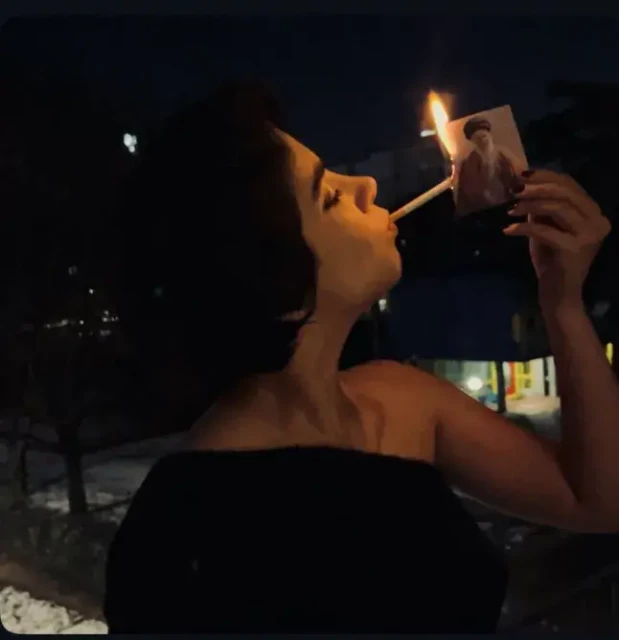 a woman lighting a cigarette with a burning phot of the Ayatollah