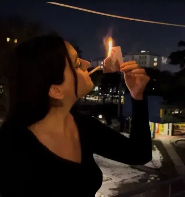 a woman lighting a cigarette with a burning phot of the Ayatollah