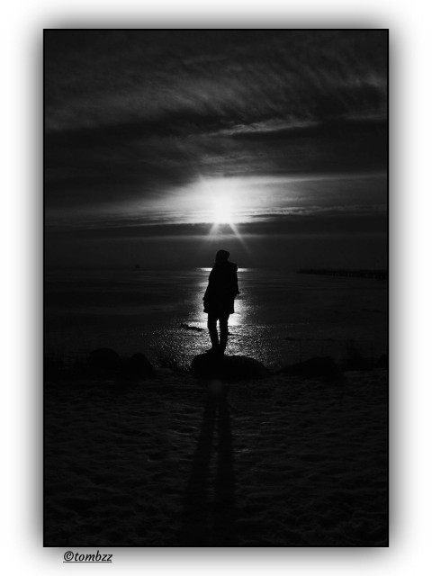 Black-and-white photo in deep shades of grey, full of contrast and dramatic light. A woman stands on the rocky edge of a frozen bay, her silhouette sharp against the setting sun. A beam of sunlight stretches across the ice, reaching the shore and bouncing off the cracks and textures in the frozen surface. On the right side of the frame there’s a pier, dark and almost blending into the shadows. The sky is striking, packed with layered clouds that seem to move and shift. The whole scene feels cold and quiet. It’s that moment right before darkness takes over, when the light is still holding on. The mood is one of solitude, focus and something hard to name that’s just about to end.
