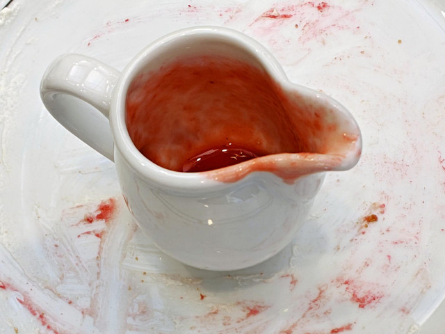 A photo showing a white ceramic creamer containing the dregs of a bright red liquid. It sits on a plate smeared with traces of red sauce.