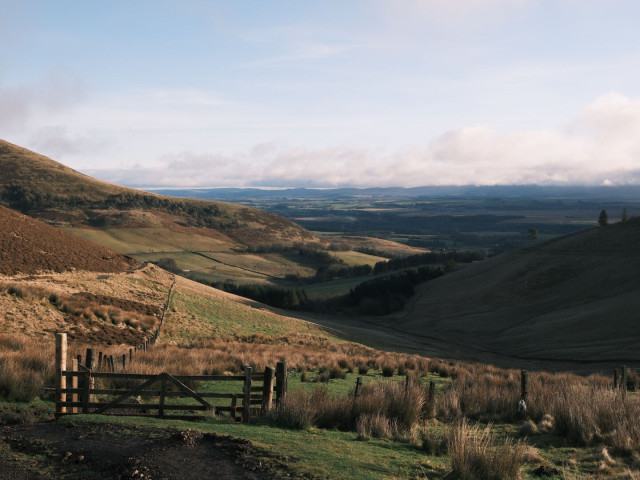 Moorland and hill farm landscape, dominated by heather, bracken and rough grazing. Bright, low sun makes a hazy sky and deep shadows. A wooden farm gate in the foreground leads into a weaving glen and on to the low ground.