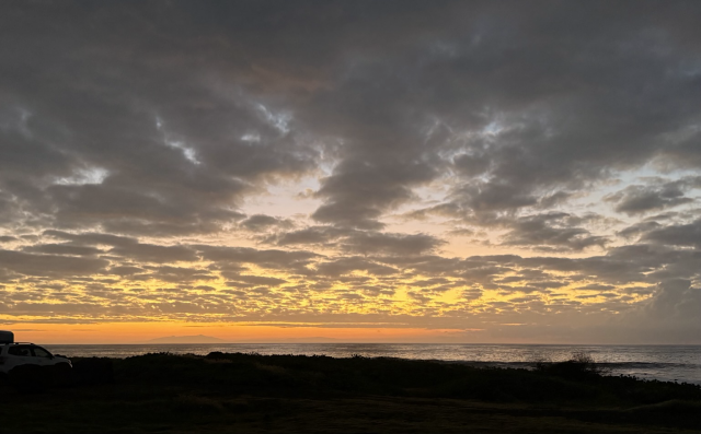 Sky over the ocean as dawn lights up the sky. Puffy, broken clouds all over the sky. Orange and yellow at the horizon, fading to pink, purple and lightest blue behind the clouds.