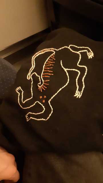 A black shirt with a four-legged creature playfully outlined in white embroidery. The creature has its small head thrown upward, and its limbs are wavy as if dancing. Its tail is bent across its back, almost touching the uplifted chin. There are orange stripes embroidered along its back and down its long tail, and three orange spots on its chest.